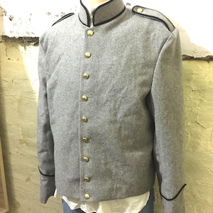 Könnte beinhalten: Graue Wolljacke mit Stehkragen und goldenen Knöpfen. Die Jacke hat schwarze Paspeln am Kragen, an den Manschetten und an den Schulterklappen. Die Jacke wird auf einer Schaufensterpuppe präsentiert, darunter ein weißes Hemd und blaue Hosen.