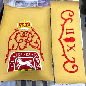 Może przedstawiać: Dwa prostokątne kawałki tkaniny w kolorze musztardowym z czerwonymi i złotymi haftami. Jeden przedstawia koronę, ozdobny skrypt i herb z białym koniem. Drugi ma dekoracyjny wzór z rzymskimi cyframi i stylizowanym płomieniem.