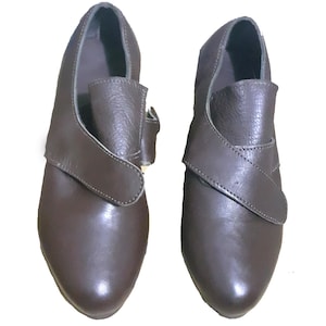 May include: A pair of brown leather shoes. Each shoe has a distinctive strap design that crosses over the top and fastens with a buckle. The shoes have a rounded toe and a low heel, suitable for various occasions.