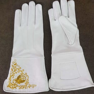 May include: A pair of white leather gloves with long cuffs. The left glove features gold embroidery of a bird and floral design. The gloves are likely for protection or decorative purposes.