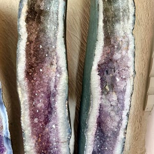 Op de afbeelding: Twee grote amethistgeodes met een donkergroene buitenkant en een sprankelend interieur van paarse en witte kristallen. De geodes worden rechtop gepresenteerd en tonen hun natuurlijke schoonheid en unieke formaties.