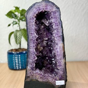 Può includere: Una grande geode di ametista verticale caratterizzata da strutture cristalline viola intenso all'interno di un centro cavo. L'esterno è racchiuso in una base scura e solida con una piccola etichetta bianca che riporta il testo PESO 21.72 kg.