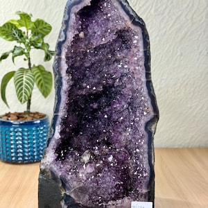 Pode incluir: Uma geode de ametista alta e vertical com um interior cristalino roxo profundo, apresentando pontas cintilantes e irregulares. O exterior é uma estrutura de pedra escura e rugosa com uma base plana. Um pequeno adesivo branco está colado.