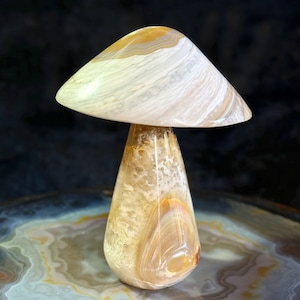 Pode incluir: Uma escultura de pedra polida em forma de cogumelo, com um chapéu cónico largo com padrões de faixas em creme, bege e branco. O caule é uma pedra cónica de cor âmbar translúcida com texturas cristalinas e faixas internas visíveis.