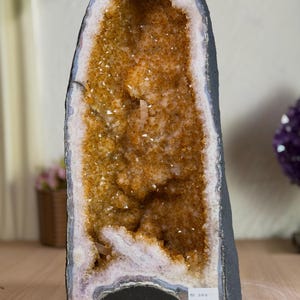 May include: A tall, vertical citrine geode featuring a hollow center filled with sparkling, golden-yellow quartz crystals. The exterior is a dark, rough, natural stone crust, while the inner rim displays layers of white and grey agate banding.
