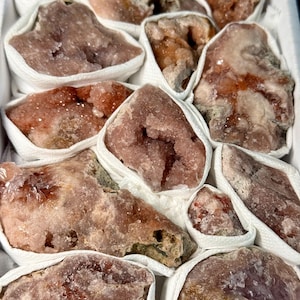 May include: A collection of raw, unpolished mineral specimens in shades of pink, peach, and rust, each individually wrapped in white paper. The stones feature crystalline textures, jagged edges, and varied geological formations packed together.