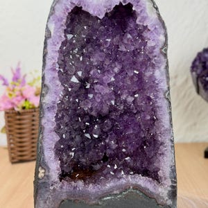 Könnte beinhalten: Eine hohe, aufrecht stehende Amethyst-Geode mit einem tiefvioletten, kristallinen Innenraum, der von einer dicken, dunkelgrauen natürlichen Gesteinsschicht umgeben ist. Die Kristallformation zeigt verschiedene Violetttöne.