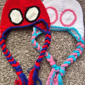 May include: Two handmade crochet hats. One is red with black and white eye details and blue trim, the other is white with pink and blue eye details and blue trim. Both have braided earflaps with tassels.