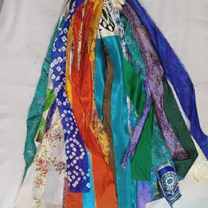 Könnte beinhalten: Ein farbenfroher Quastenanhänger aus Stoffstreifen in verschiedenen Farben, darunter Blau, Orange, Grün und Lila. Die Stoffstreifen haben unterschiedliche Muster und Texturen, wodurch ein Bohème- oder eklektischer Look entsteht. Der Quastenanhänger dient wahrscheinlich der Dekoration.