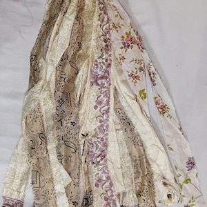 Op de afbeelding: Een verzameling stoffen stroken met diverse patronen, waaronder bloemenprints in roze en groen, gedetailleerde zwarte paisleyontwerpen op een beige achtergrond en effen crèmekleurig kant, allemaal bovenaan samengebonden met gerafelde randen.