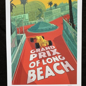 May include: A yellow race car speeds around a red track with the words "Grand Prix of Long Beach" in white letters. The track is surrounded by palm trees and buildings. The words "Bobba Toyota" are visible on a building in the background.