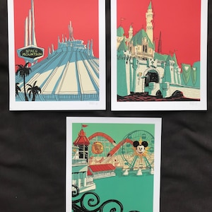 May include: Three framed art prints with illustrations of amusement park attractions. One shows Space Mountain, another a castle, and the third a roller coaster and Ferris wheel. The prints have a red, teal, and white colour scheme.