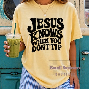 Op de afbeelding: Een lichtgeel t-shirt met de zwarte tekst "JESUS KNOWS WHEN YOU DON'T TIP" in een retro lettertype. Het shirt wordt gecombineerd met blauw-wit gestreepte shorts. Een doorzichtige plastic beker met een groene drank wordt in de hand gehouden.