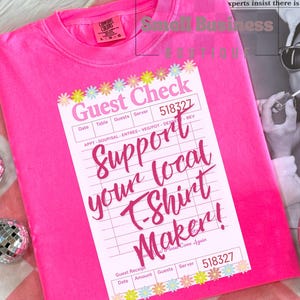 May include: Bright pink t-shirt with a printed "Guest Check" design. The design includes the text "Support your local T-Shirt Maker!" and a floral border. The shirt has a crew neck and short sleeves.