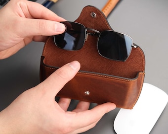 May include: Sunglasses with dark lenses and a silver frame are held in a brown leather case. The case has a snap closure and a curved top. A white computer mouse is in the background.