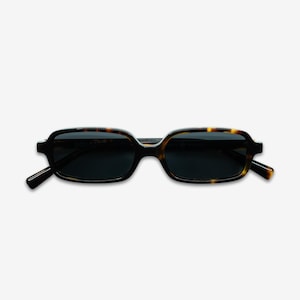 May include: Rectangular sunglasses with a tortoiseshell pattern frame and dark lenses. The sunglasses have a classic design with a slightly rounded shape. The temples are black. The brand name is printed on the left lens.