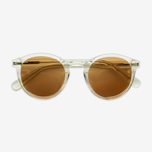 May include: Clear-framed sunglasses with round lenses in a warm brown hue. The sunglasses have a classic design with a transparent frame and temples. The lenses are a solid color, providing a stylish and functional accessory.