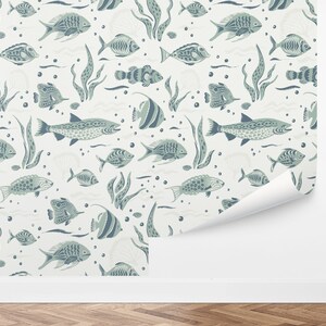 May include: Wallpaper featuring a repeating pattern of stylized fish, seaweed, and bubbles in shades of blue and gray on a cream background. The design includes various fish species and aquatic elements, creating a nautical theme. A section of the wallpaper is peeled back.