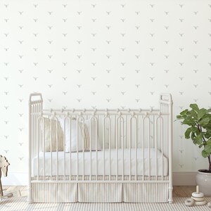 May include: A white crib with a white mattress and a white crib skirt. The crib is in a room with white walls and a gray patterned wallpaper. There is a green plant in a pot on the right side of the image.