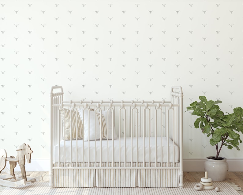 May include: A white crib with a white mattress and a white crib skirt. The crib is in a room with white walls and a gray patterned wallpaper. There is a green plant in a pot on the right side of the image.