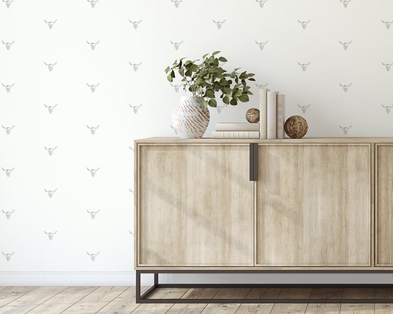 May include: A light wood cabinet with two doors and black metal legs. The cabinet is decorated with a vase of green plants and a stack of books. The wall behind the cabinet is white and has a pattern of small, gray bull skulls.