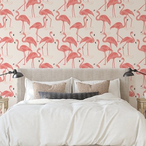 Op de afbeelding: Een slaapkamer met een wit bed en roze flamingo behang. Het behang heeft een herhalend patroon van roze flamingo's die in een waterpartij staan. Het bed heeft een wit dekbed en twee kussens. Er staan twee nachtkastjes met lampen aan weerszijden van het bed.