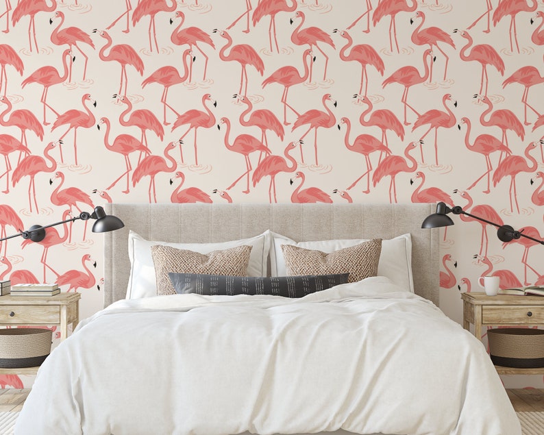 Op de afbeelding: Een slaapkamer met een wit bed en roze flamingo behang. Het behang heeft een herhalend patroon van roze flamingo's die in een waterpartij staan. Het bed heeft een wit dekbed en twee kussens. Er staan twee nachtkastjes met lampen aan weerszijden van het bed.