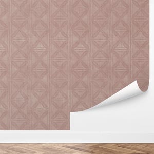 May include: A roll of wallpaper with a repeating geometric pattern in a muted rose color. The design features a series of connected diamond shapes. A corner of the wallpaper is peeled back, revealing a white wall. The wallpaper is on a wall above a wooden floor.