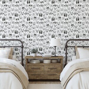 Pode incluir: Duas camas de solteiro com roupa de cama branca e cobertores bege em um quarto com paredes brancas e papel de parede com rostos de c&atilde;es em preto e branco. Uma mesa de cabeceira de madeira com uma l&acirc;mpada e livros fica entre as camas.