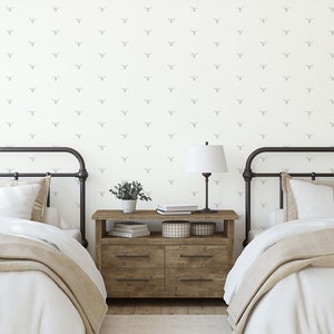 May include: Two twin beds with white bedding and tan blankets in a bedroom with white walls featuring a gray and white patterned wallpaper with a repeating design of a stylized bull's head. A wooden nightstand with two drawers sits between the beds with a lamp, books, and a potted plant on top.