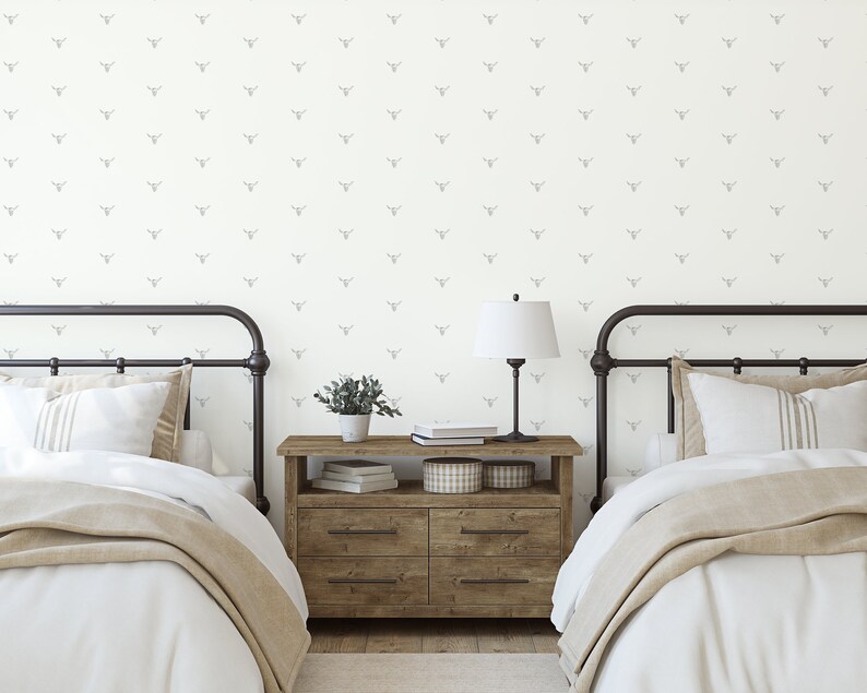 May include: Two twin beds with white bedding and tan blankets in a bedroom with white walls featuring a gray and white patterned wallpaper with a repeating design of a stylized bull's head. A wooden nightstand with two drawers sits between the beds with a lamp, books, and a potted plant on top.