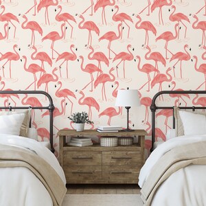 Op de afbeelding: Twee eenpersoonsbedden met witte beddengoed en roze flamingo behang. De bedden hebben zwarte metalen frames en staan in een kamer met een houten nachtkastje en een lamp.