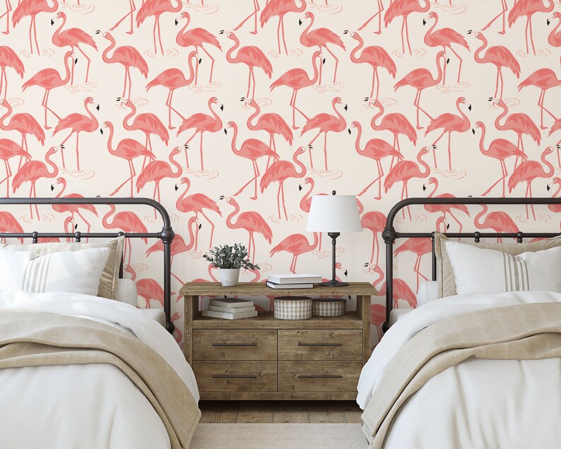 Op de afbeelding: Twee eenpersoonsbedden met witte beddengoed en roze flamingo behang. De bedden hebben zwarte metalen frames en staan in een kamer met een houten nachtkastje en een lamp.