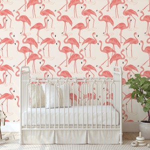 Op de afbeelding: Een wit babybedje met een roze flamingo behangpatroon. Het babybedje heeft een wit matras en een wit schommelpaard ervoor. Rechts van het babybedje staat een groene plant in een witte pot.