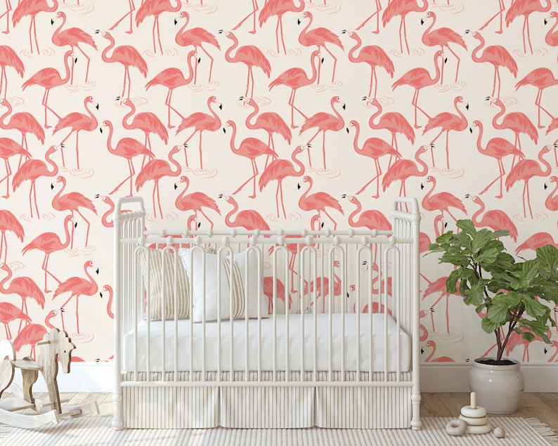 Op de afbeelding: Een wit babybedje met een roze flamingo behangpatroon. Het babybedje heeft een wit matras en een wit schommelpaard ervoor. Rechts van het babybedje staat een groene plant in een witte pot.