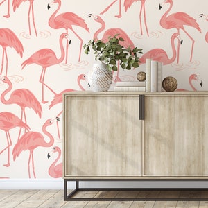 Op de afbeelding: Een licht houten kast met twee deuren tegen een witte achtergrond met een roze flamingo-patroon. De kast heeft zwarte metalen poten en grepen. Een plant in een pot staat bovenop de kast.