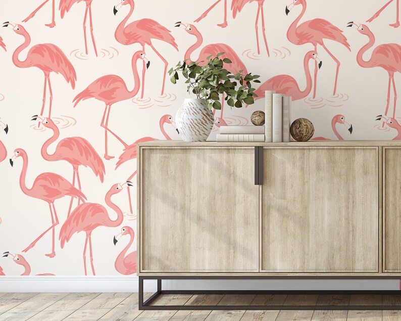 Op de afbeelding: Een licht houten kast met twee deuren tegen een witte achtergrond met een roze flamingo-patroon. De kast heeft zwarte metalen poten en grepen. Een plant in een pot staat bovenop de kast.