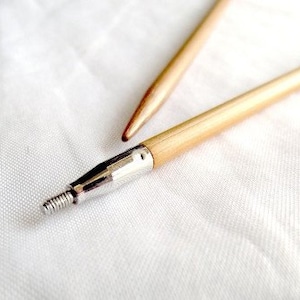 Peut inclure: Deux aiguilles à tricoter en bois avec des pointes métalliques. Les aiguilles sont posées sur une surface blanche.