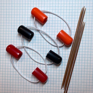 Op de afbeelding: Een set van vijf breinaalden met oranje, zwarte en rode eindkappen. De naalden zijn met een wit elastiekje bij elkaar gehouden.