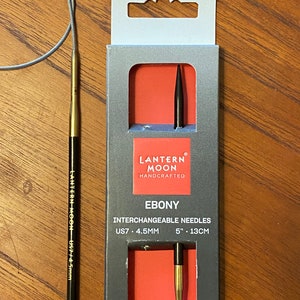 May include: A pair of black and gold interchangeable knitting needles in a clear plastic package. The package is labeled "Lantern Moon Handcrafted Ebony" and includes the size US7/4.5mm and 5"/13cm.