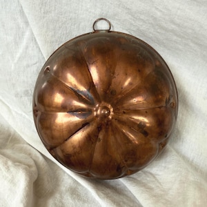 Puede incluir: Un molde de cobre vintage con forma de calabaza. El molde tiene una forma redonda con estrías verticales y un pequeño anillo para colgar. El cobre tiene una pátina cálida y envejecida.