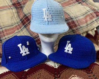 May include: Three crocheted baseball caps in shades of blue. The caps feature the white letters "LA" on the front. One cap is light blue, and the other two are a darker blue. Some caps have pearl accents.