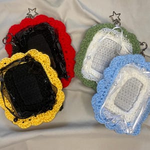May include: Four crocheted phone cases in red, yellow, green, and blue. Each case has a scalloped edge and a star-shaped charm. The cases feature black fabric and decorative elements like pearls and lace.
