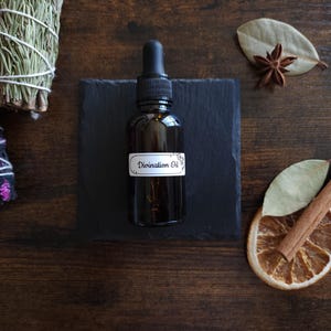 May include: A dark glass bottle with a dropper, labeled "Divination Oil," sits on a black square. The bottle is surrounded by dried herbs, spices, and a dried orange slice, all arranged on a dark wooden surface.