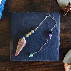 May include: A pendulum with a faceted, reddish-brown crystal point and a silver chain. The chain is adorned with colorful beads and a star and crescent moon charm. The pendulum is designed for divination and spiritual practices.
