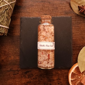 May include: A clear glass bottle filled with pink salt and a cork stopper. The label reads "Witch's Fire Salt". The bottle sits on a black square, surrounded by dried herbs, spices, and a dried orange slice on a wooden surface.