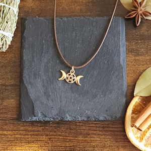 May include: A gold-colored pendant necklace featuring a triple moon and pentacle charm. The necklace is displayed on a dark gray square stone, with dried herbs, spices, and a dried orange slice in the background.
