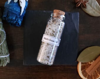 May include: A clear glass bottle filled with green salt, labeled "Witch's Green Salt", sits on a black surface. The bottle is corked and surrounded by dried herbs and spices, including sage, bay leaves, and star anise, creating a rustic, natural aesthetic.