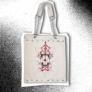 Könnte beinhalten: Weißer Canvas-Tote-Bag mit einem roten und schwarzen Tribal-Schädel-Design. Die Tasche hat weiße Griffe mit silbernen Kettenakzenten und Metallösen am unteren Rand. Der Text "RECLAIMED BY salinkas" ist unten rechts aufgedruckt.