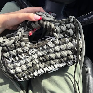 Könnte beinhalten: Eine handgestrickte Handtasche in Grau-, Weiß- und Olivgrüntönen. Die Tasche hat ein geflochtenes Design mit einem stabilen Griff und Kordelzugverschluss. Die Tasche wird in einem Auto gehalten.
